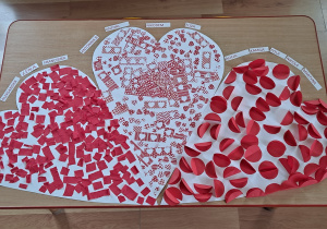 Trzy serca ozdobione przez dzieci. Na pierwszym naklejone są kawałki czerwonej bibuły. Drugie serce wykonane jest z elementów przygotowanych z wykorzystaniem dziurkacza. Na trzecim znajdują się czerwone kółka złożone na pół